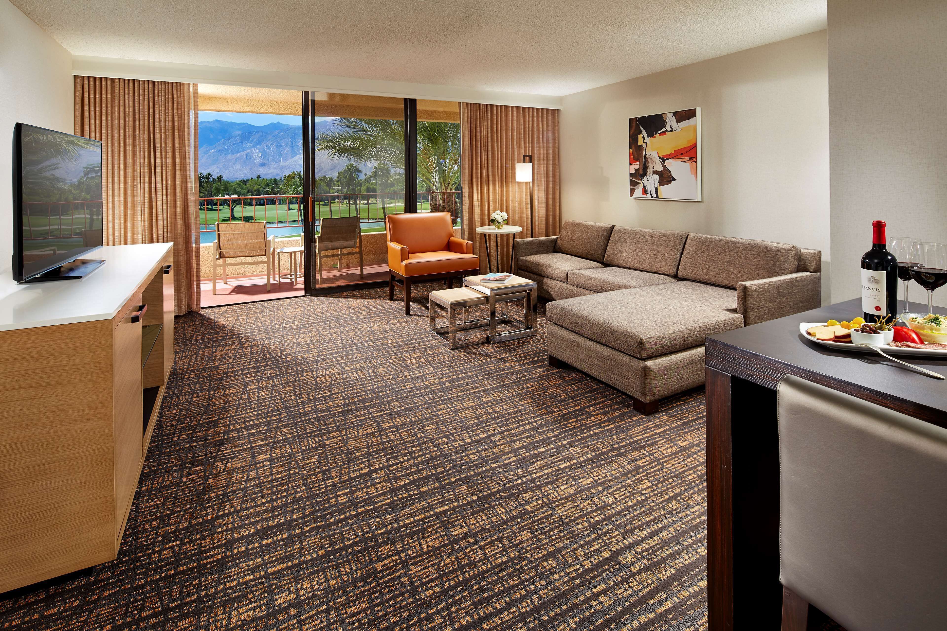A hotel room with a large window, a sofa, a chair, a small table, and a TV.