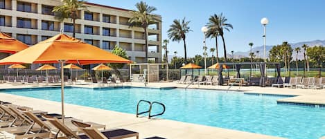A hotel pool area with sun loungers and umbrellas, a clear blue pool, and a building with balconies in the background.