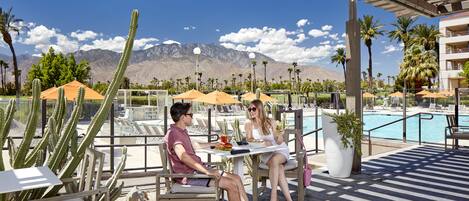 A couple dining at an outdoor table with a pool view, surrounded by cacti and palm trees.