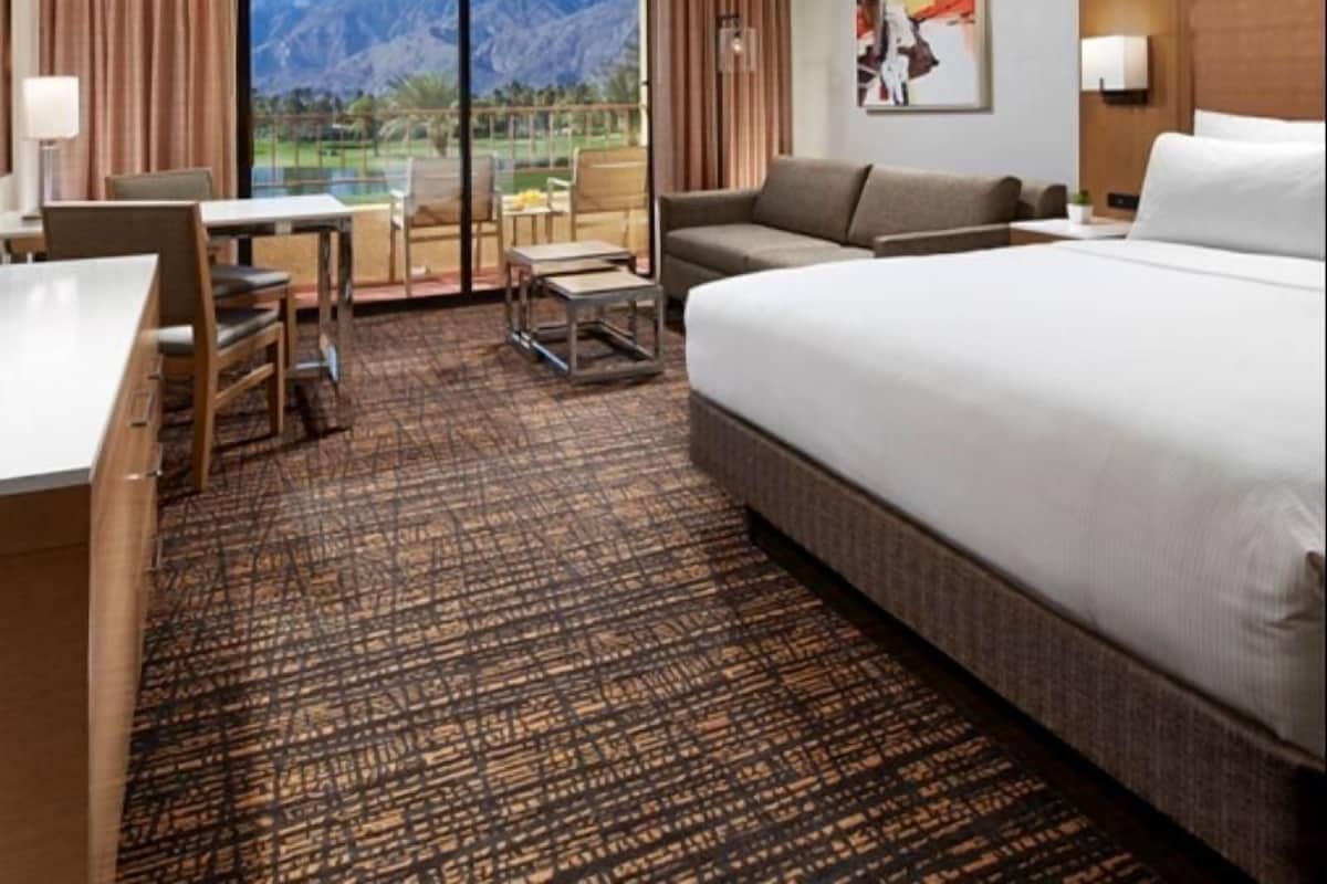 Habitación de hotel con cama, escritorio, sillón, sofá y vistas a las montañas a través de grandes ventanales.