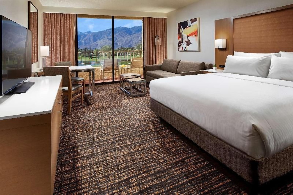 Habitación de hotel con una cama grande, un escritorio, una silla, una mesita y vistas a las montañas a través del ventanal.