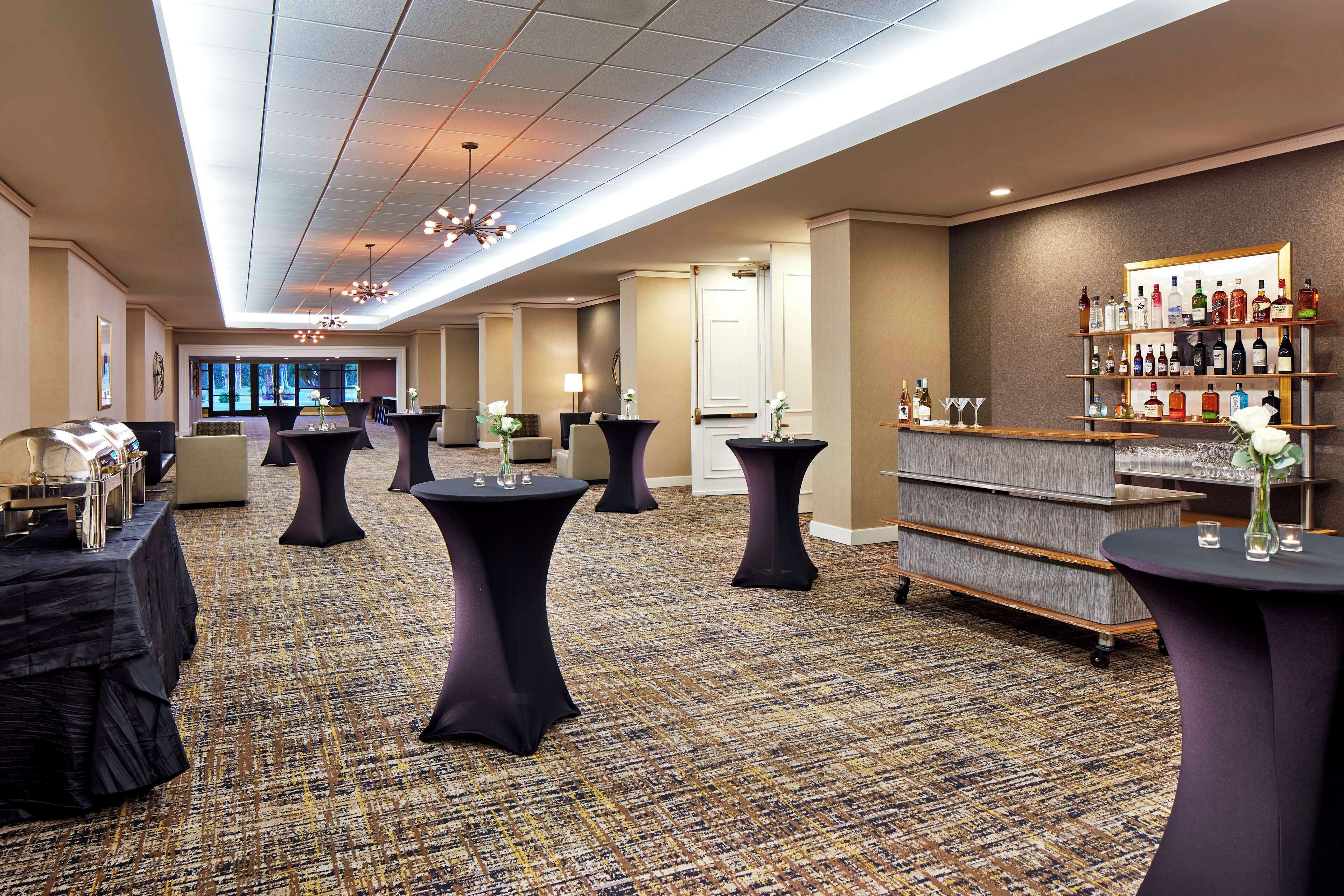 A hotel lobby with round tables, a bar area with shelves of bottles, and a carpeted floor.