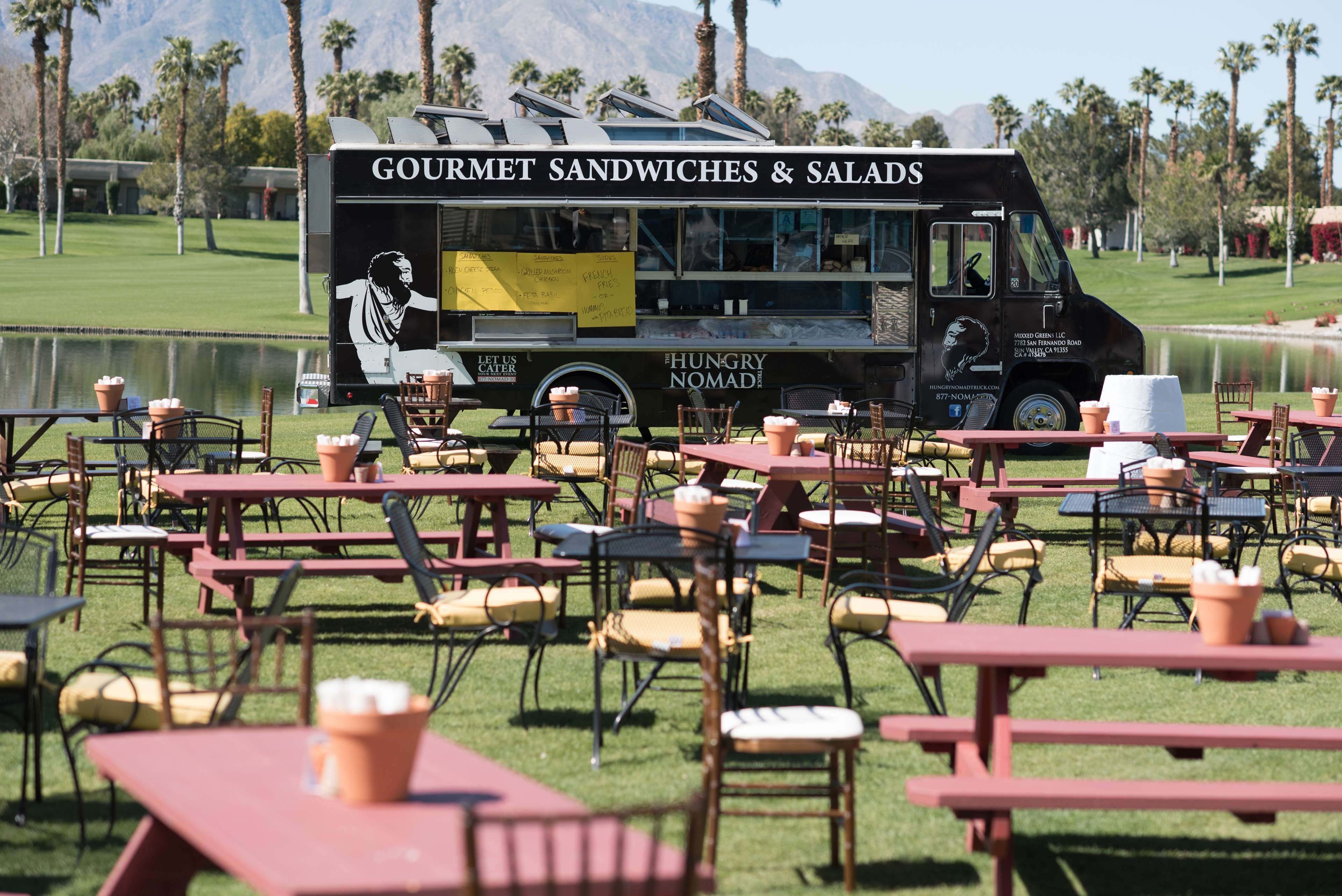 En food truck med teksten "Gourmet Sandwiches & Salads" står parkeret på en græsplæne med borde og stole, så der er mulighed for udendørs spisning.