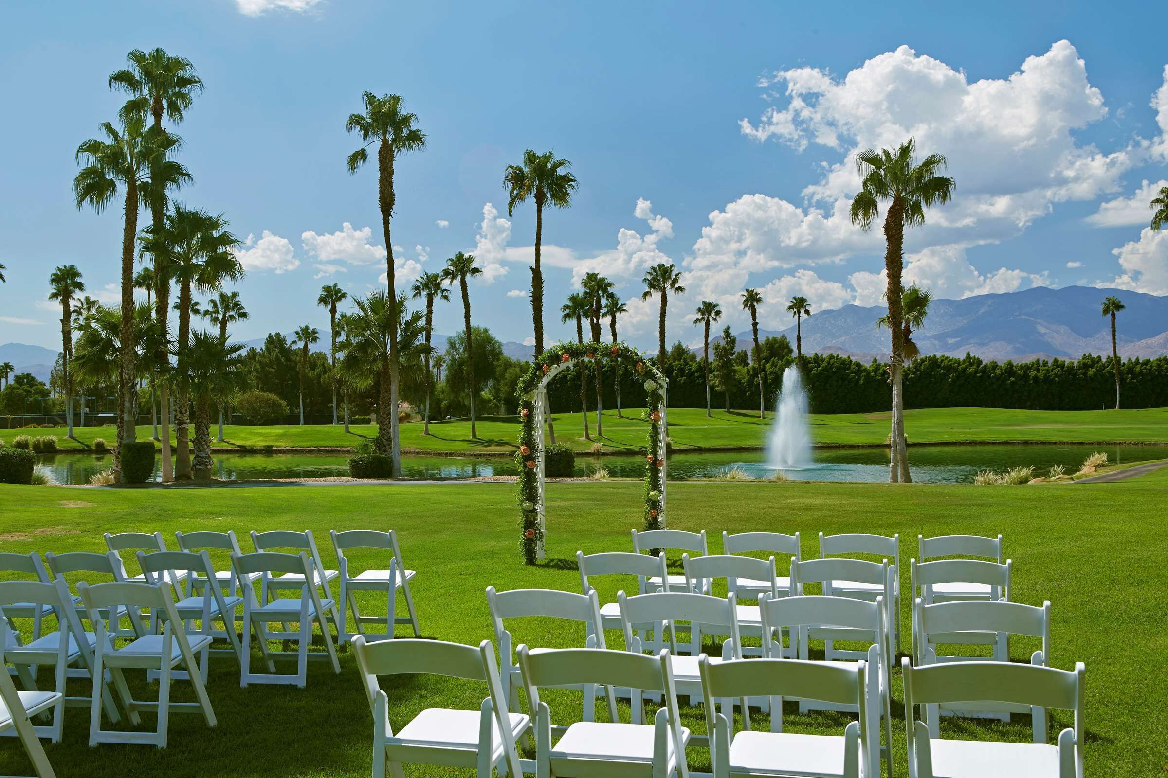 A golf course with white chairs arranged for an event, surrounded by palm trees and a fountain.