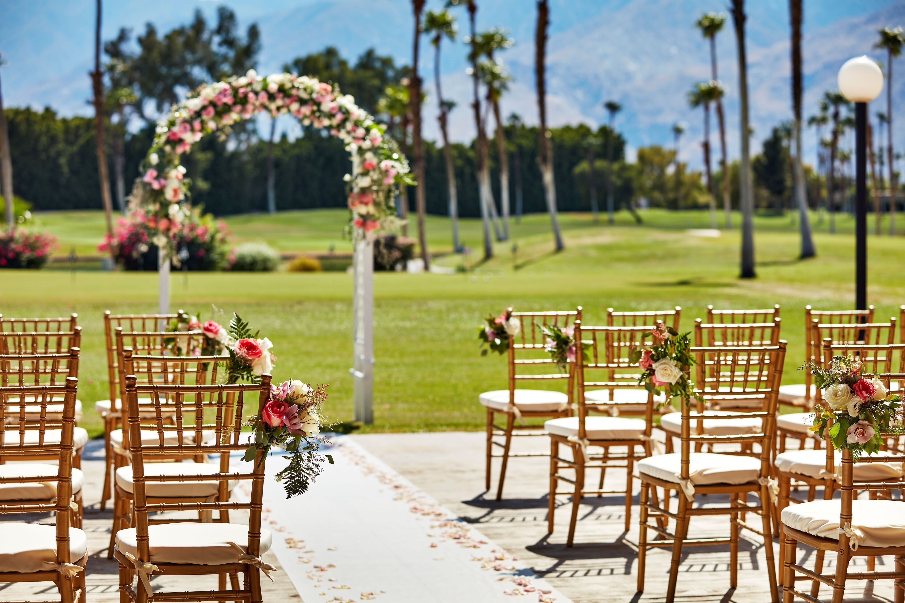 Hochzeitliche Zeremonie mit in Reihen angeordneten Stühlen, einem Blumenschmuckbogen und Blick auf einen Golfplatz im Hintergrund.