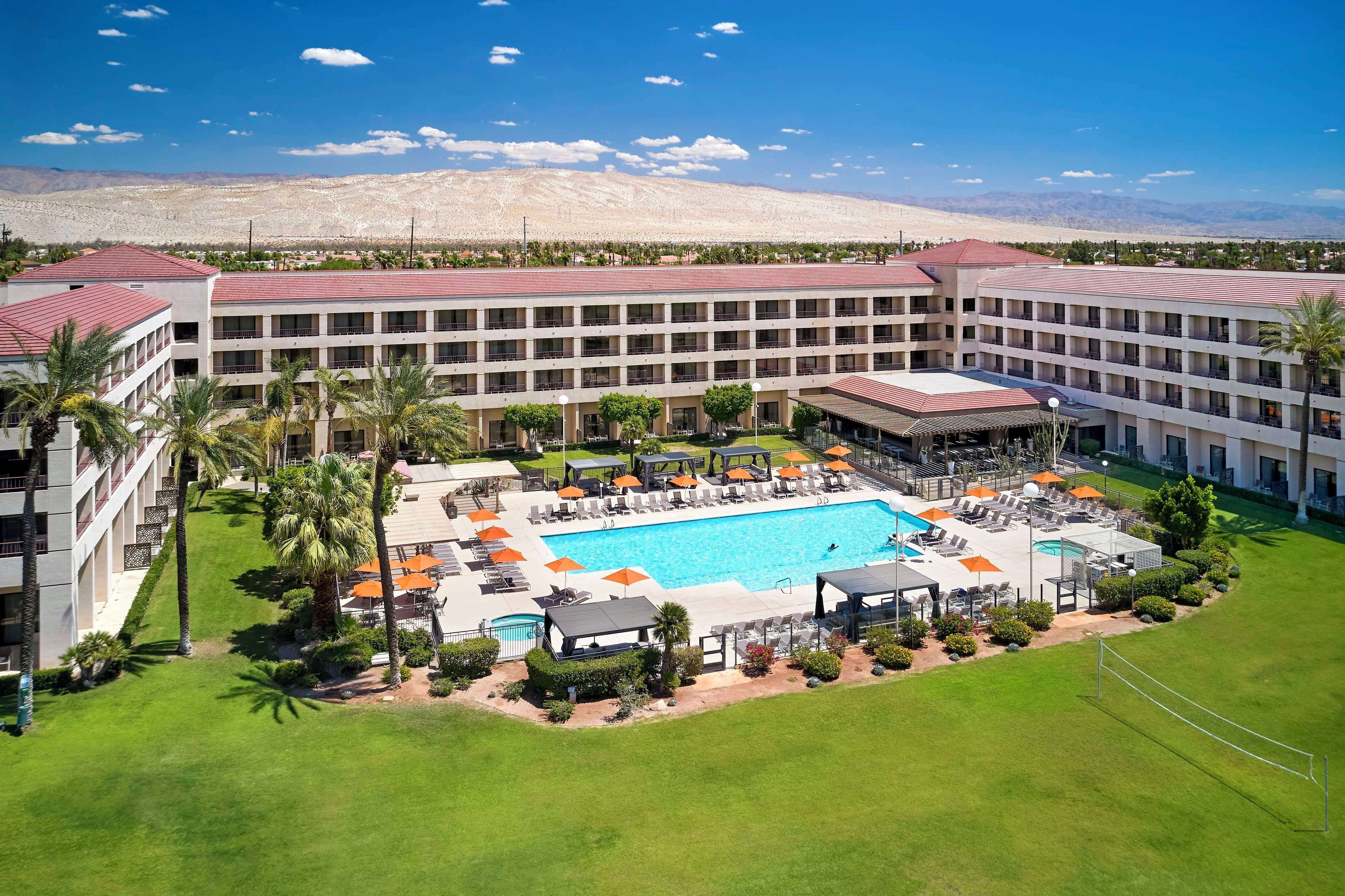 A hotel with a large swimming pool, multiple balconies, and a desert landscape in the background.