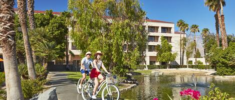 Un couple fait du vélo sur une piste aménagée, avec un élément décoratif aquatique et des palmiers en arrière-plan.