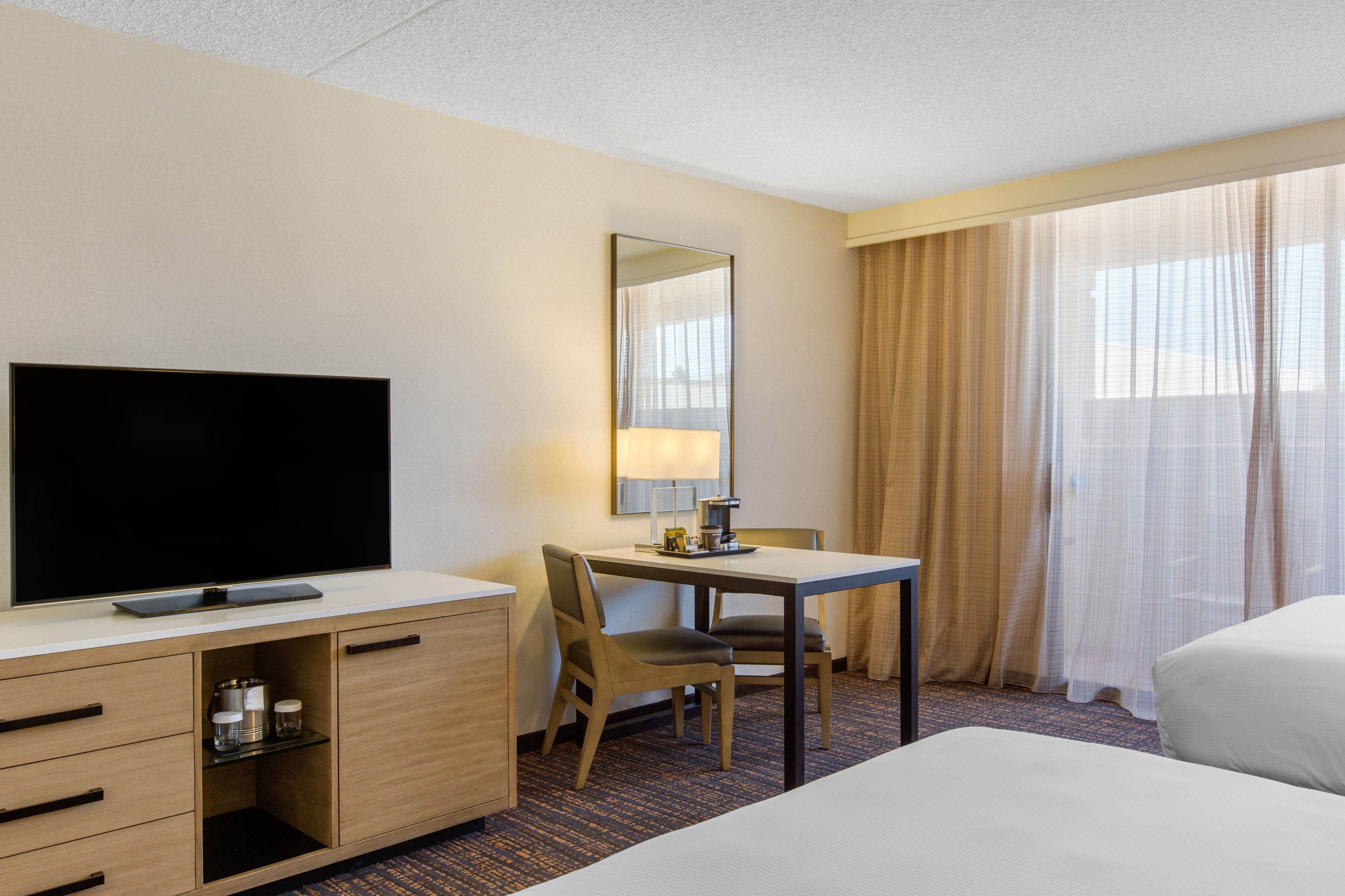 Una habitación de hotel con televisión de pantalla plana, un escritorio con lámpara, una silla, una cama y un ventanal con cortinas.