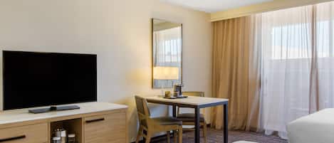 Una habitación de hotel con televisión de pantalla plana, un escritorio con lámpara, una silla, una cama y un ventanal con cortinas.