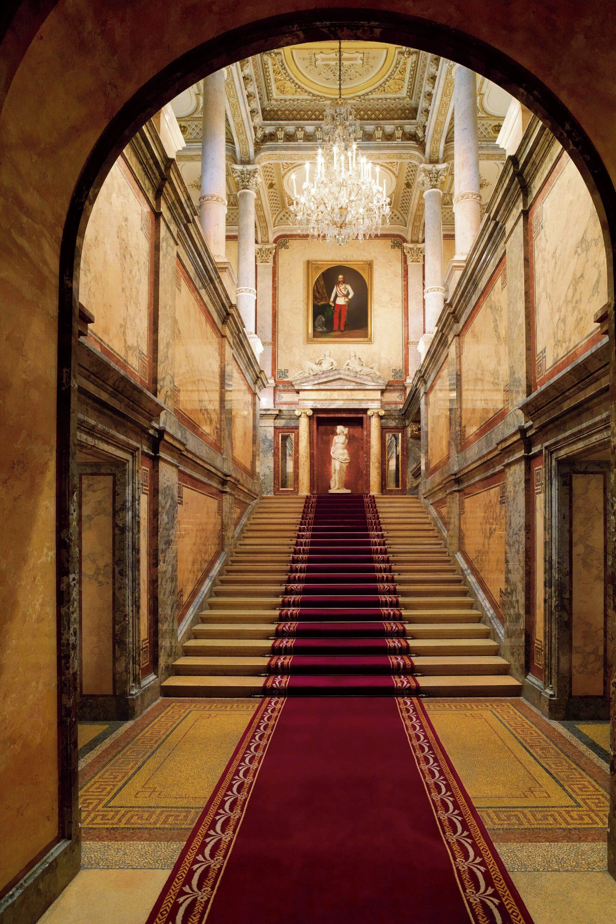 Una maestosa scalinata con un tappeto rosso che conduce a una statua in cima, affiancata da colonne e decorazioni elaborate al soffitto.