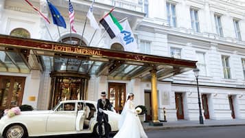 Een klassieke witte auto geparkeerd voor Hotel Imperial, met vlaggen, een statig entree en een man in pak.