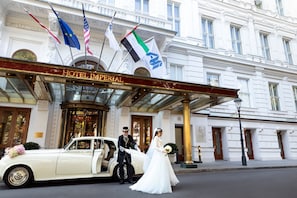 A classic white car parked in front of Hotel Imperial with flags, a grand entrance, and a man in a suit.