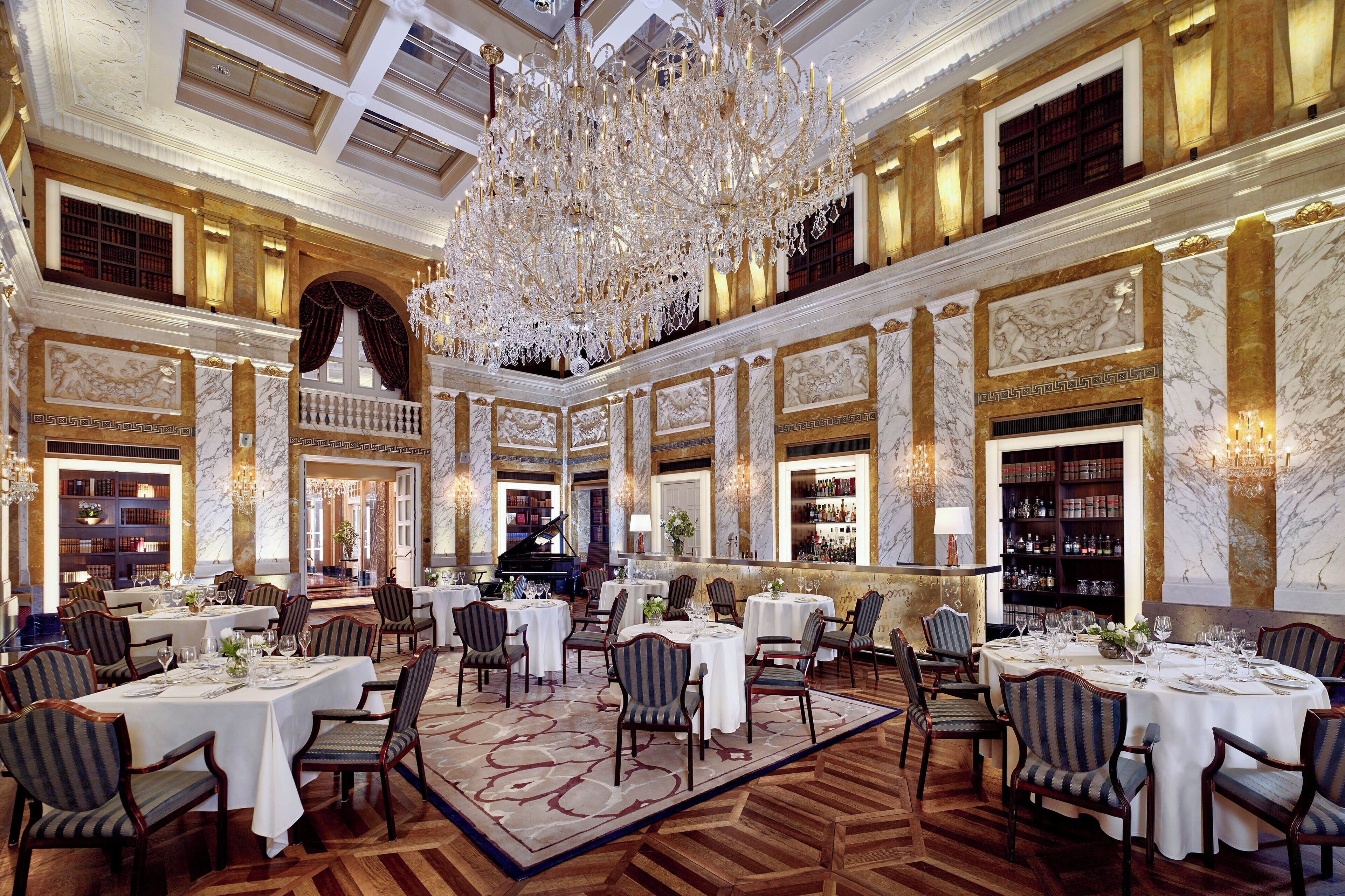 A grand dining hall with multiple tables, elegant chandeliers, marble walls, and bookshelves.