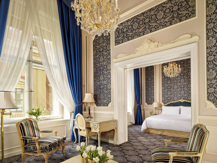 Una habitación de hotel con una cama grande, un escritorio y una silla. Se observa una araña y cortinas.