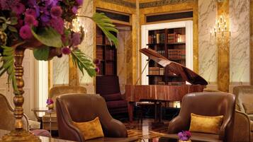 A luxurious lobby with a grand piano, leather armchairs, a marble floor, and a bookshelf filled with books.