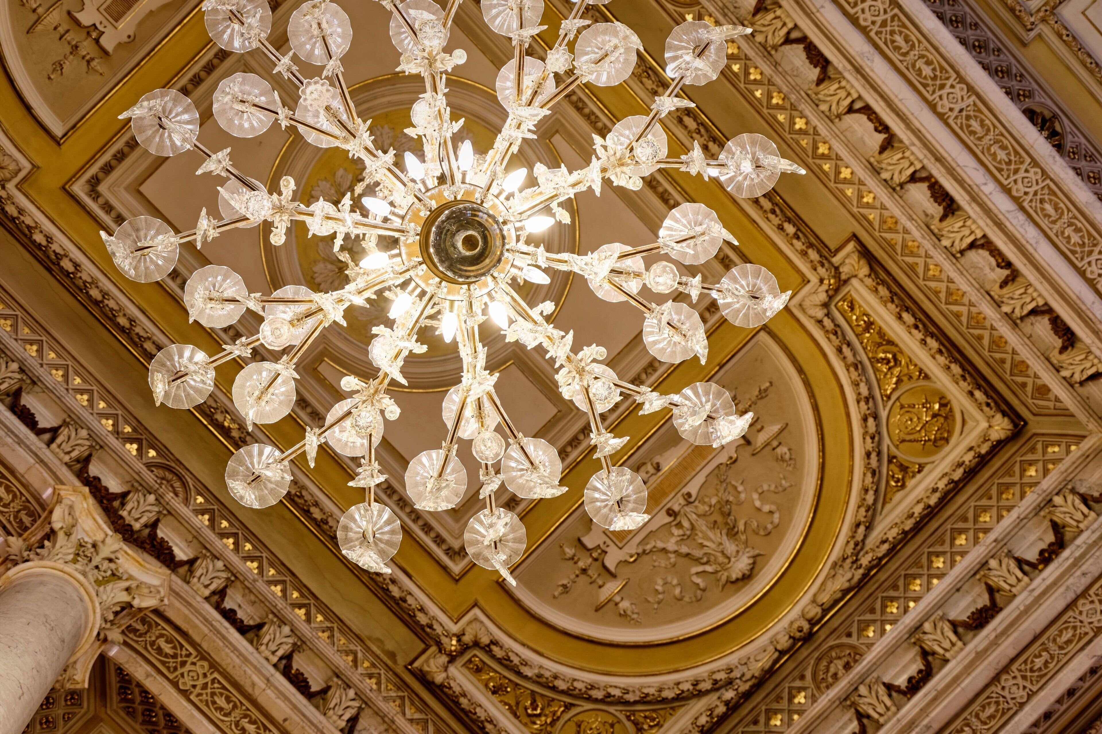 A grand chandelier with multiple arms and crystal decorations, set against a richly decorated ceiling with intricate patterns and gold accents.