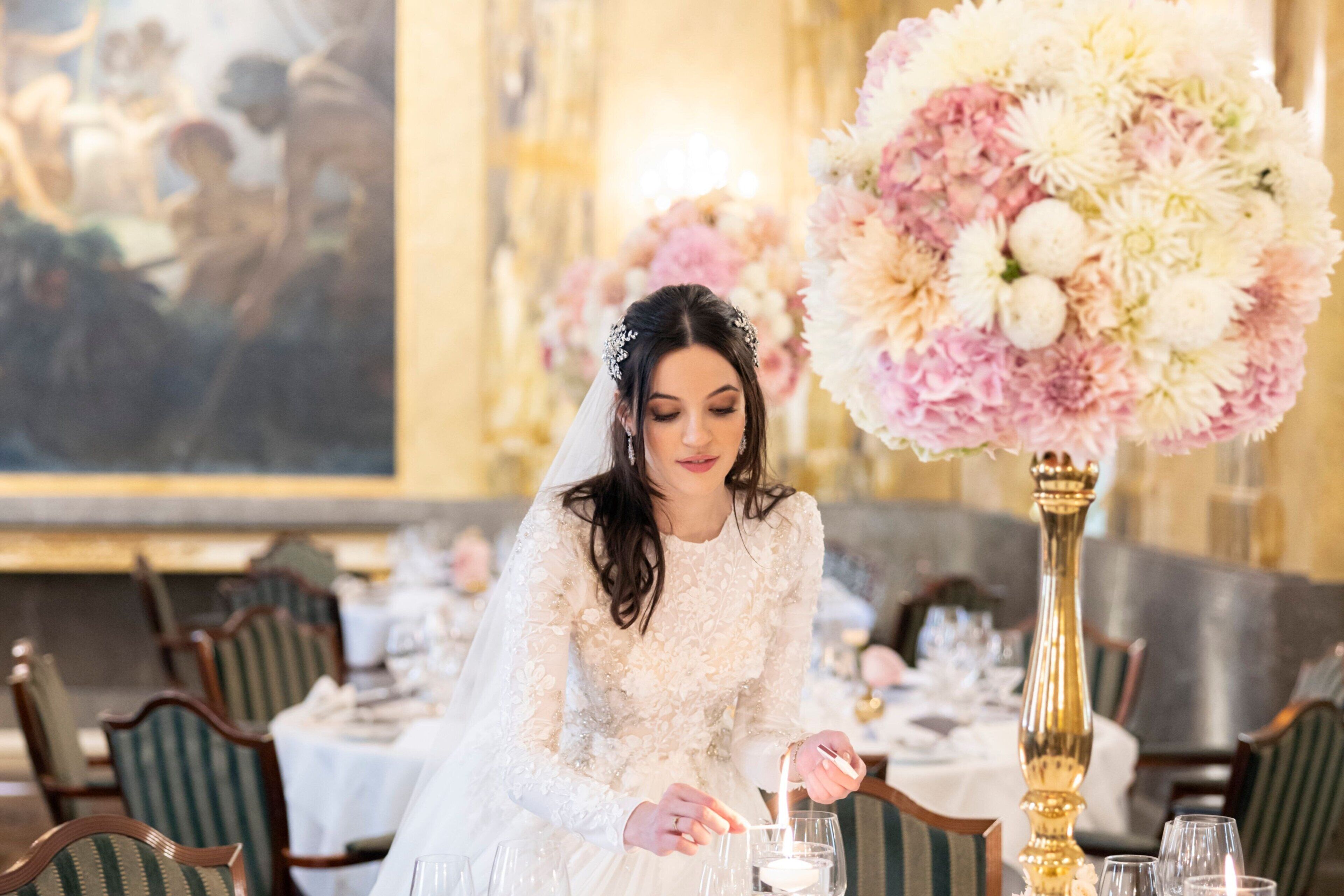 A bride in a white dress at a dining table with a large floral arrangement and a tall vase.