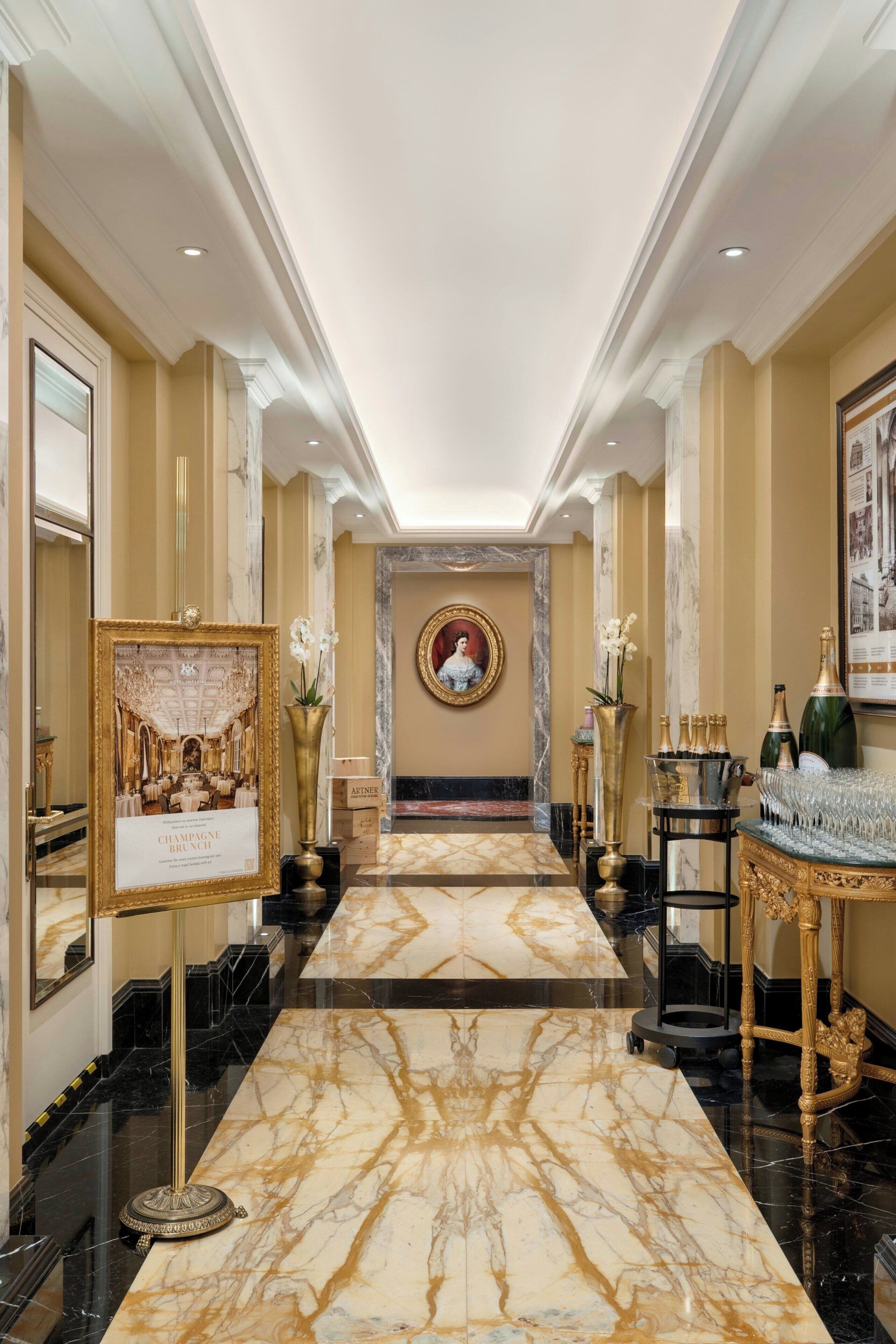 A luxurious hallway with marble flooring, framed artwork, and elegant decor including a chandelier and champagne display.