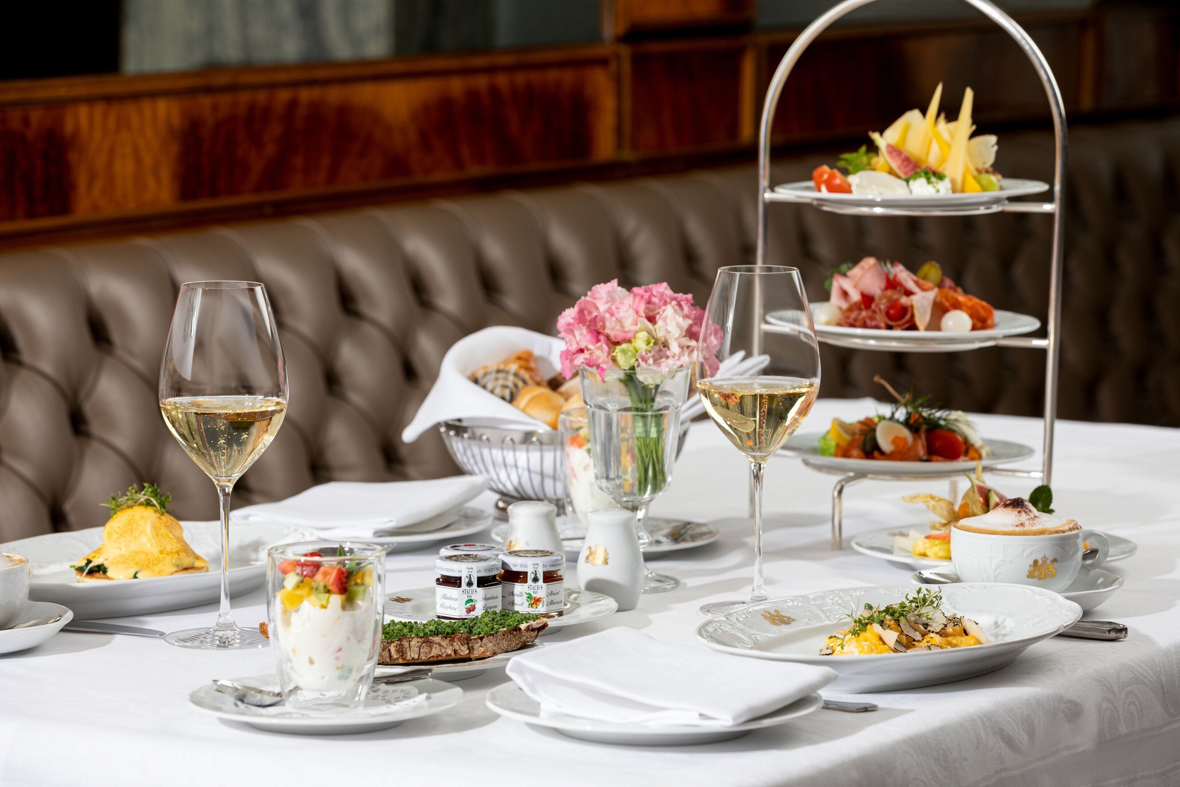 A three-tiered dessert stand with various pastries and fruits, a glass of white wine, and a bowl of flowers on a table set for a meal.