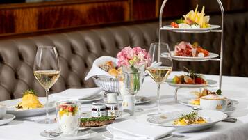 Suporte de sobremesas em três níveis com diversos pastéis e frutas, um copo de vinho branco e uma taça de flores sobre uma mesa posta para refeição.