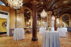A grand hall with ornate columns, chandeliers, and tables set for a formal event.