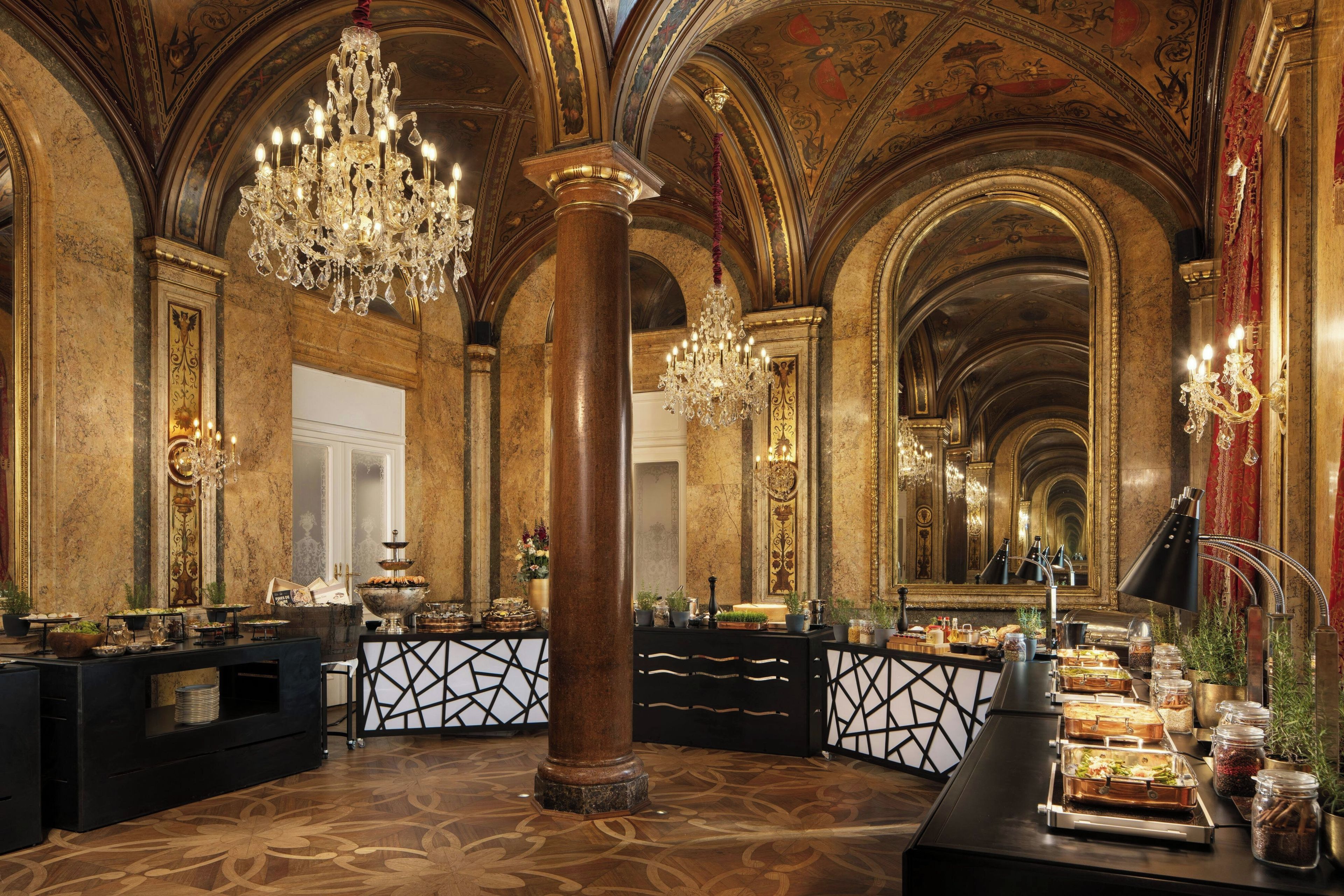 An opulent banquet hall with ornate chandeliers, marble columns, and a long buffet table.