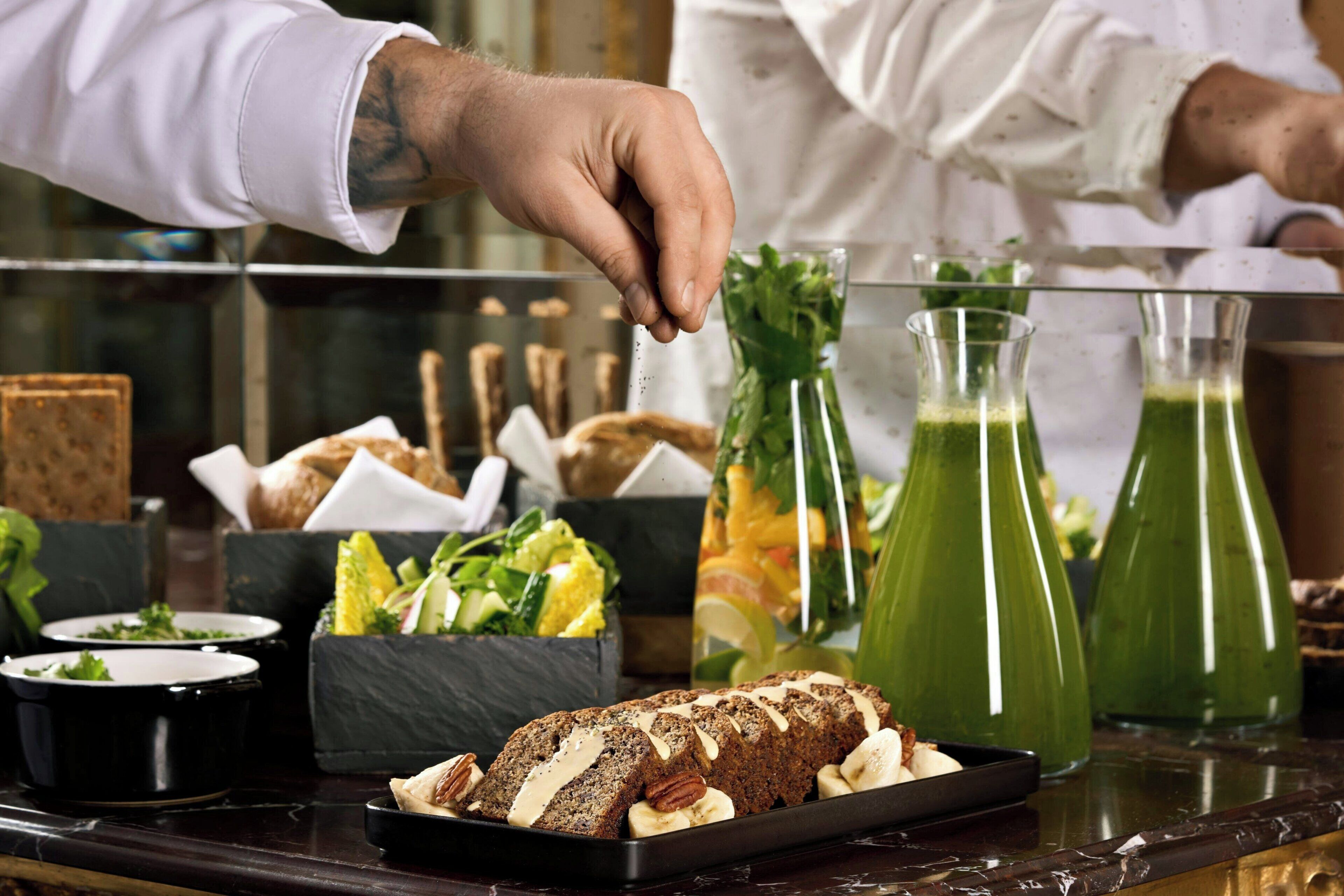 A chef's hand sprinkling seasoning on a dish, with various food items and drinks on the counter.