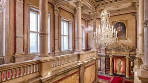 Een statige zaal met marmeren zuilen, een rood tapijt en een kroonluchter.