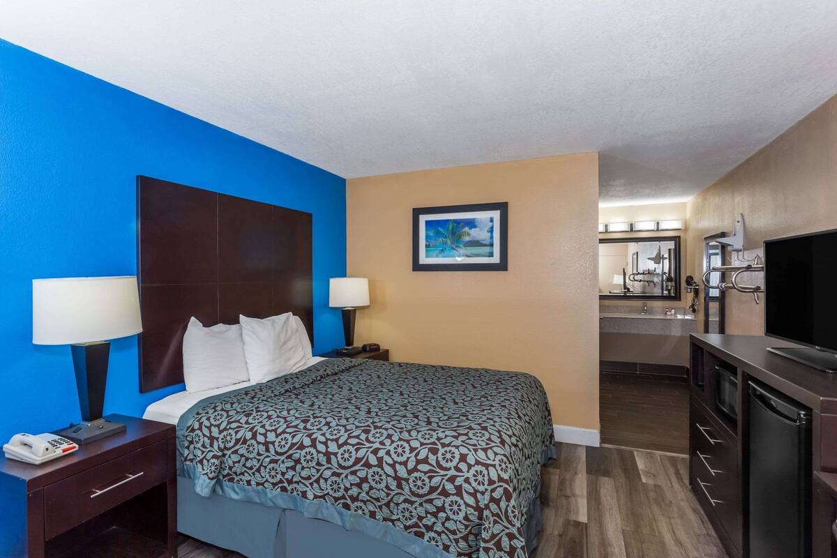 Une chambre d’hôtel comprenant un lit, des lampes de chevet, un bureau avec un téléphone, un tableau au mur, une kitchenette avec un évier et un téléviseur.