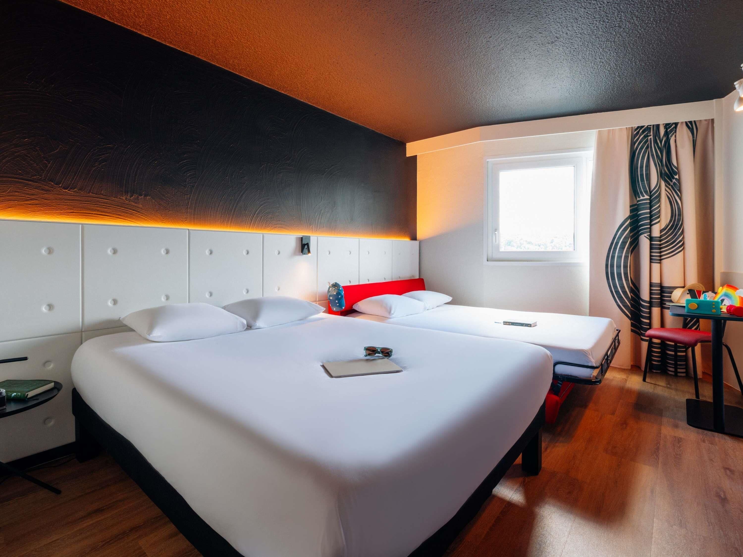 Una habitació d'hotel amb un llit gran, un altre llit més petit, un escriptori amb cadira, una finestra i vistes a l'exterior.