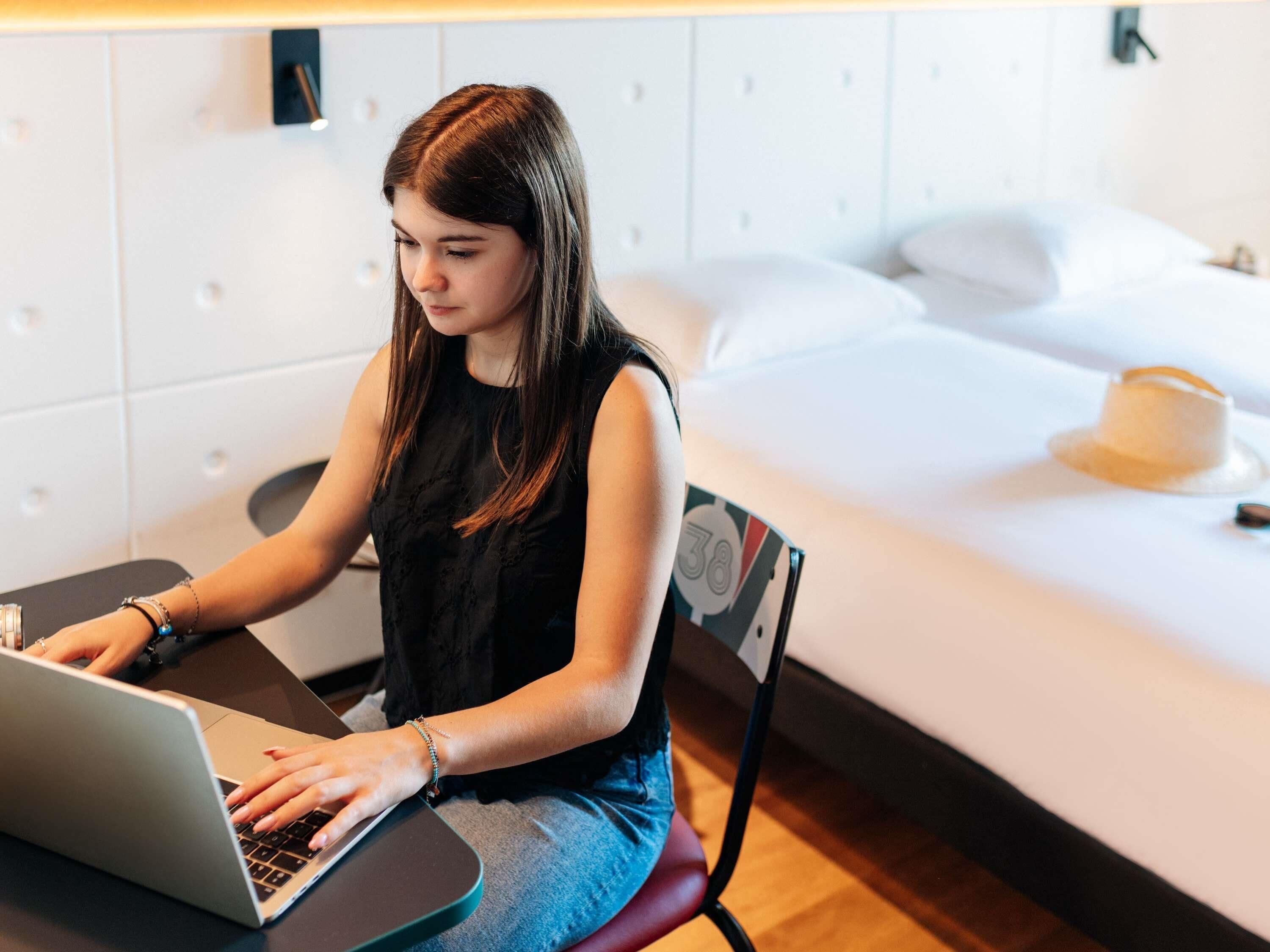 Una persona sentada en un escritorio con una computadora portátil en una habitación con una cama y una lámpara fijada a la pared.