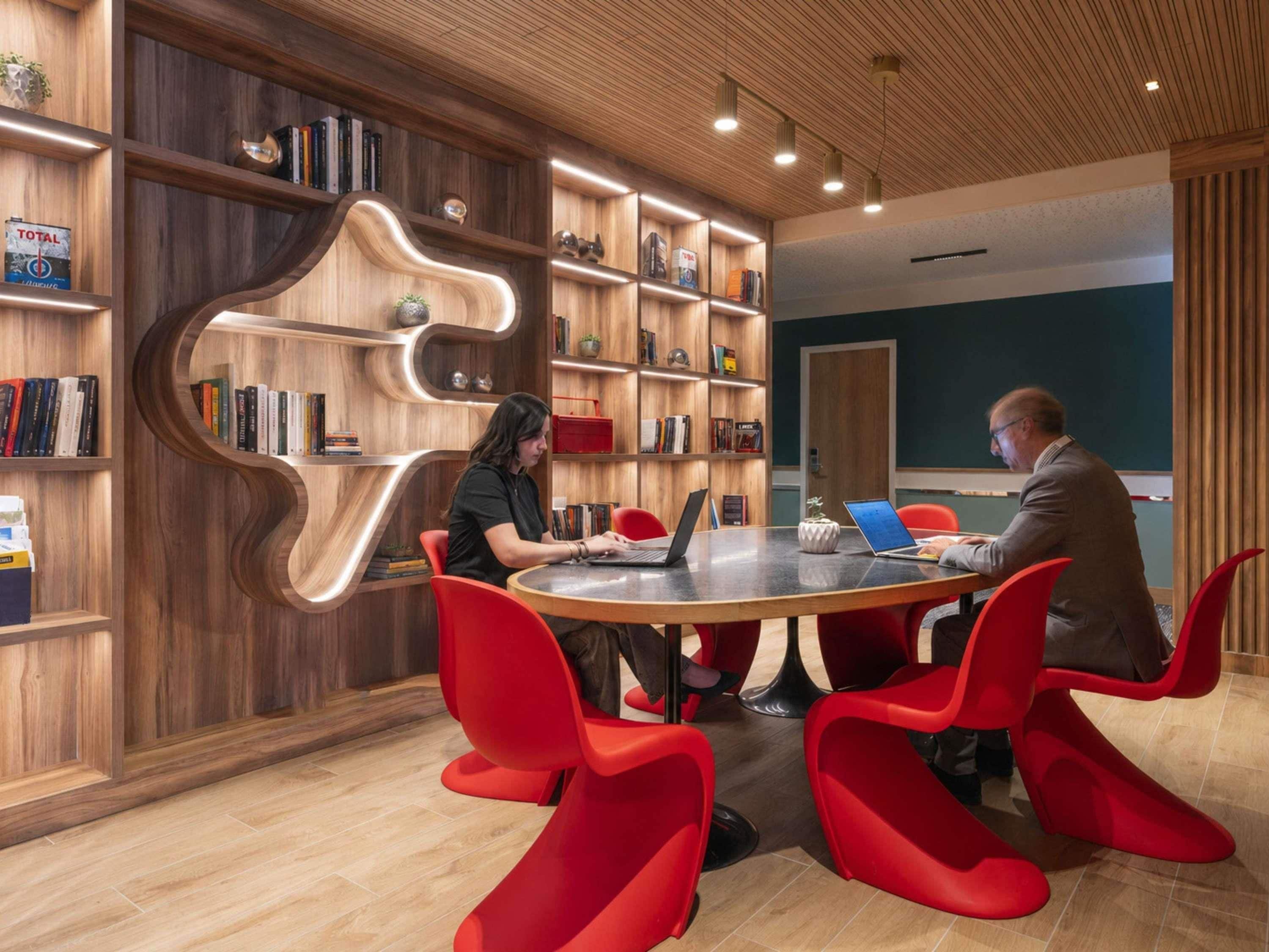 Una sala de reuniones moderna con una mesa redonda, sillas rojas y estanterías de madera.
