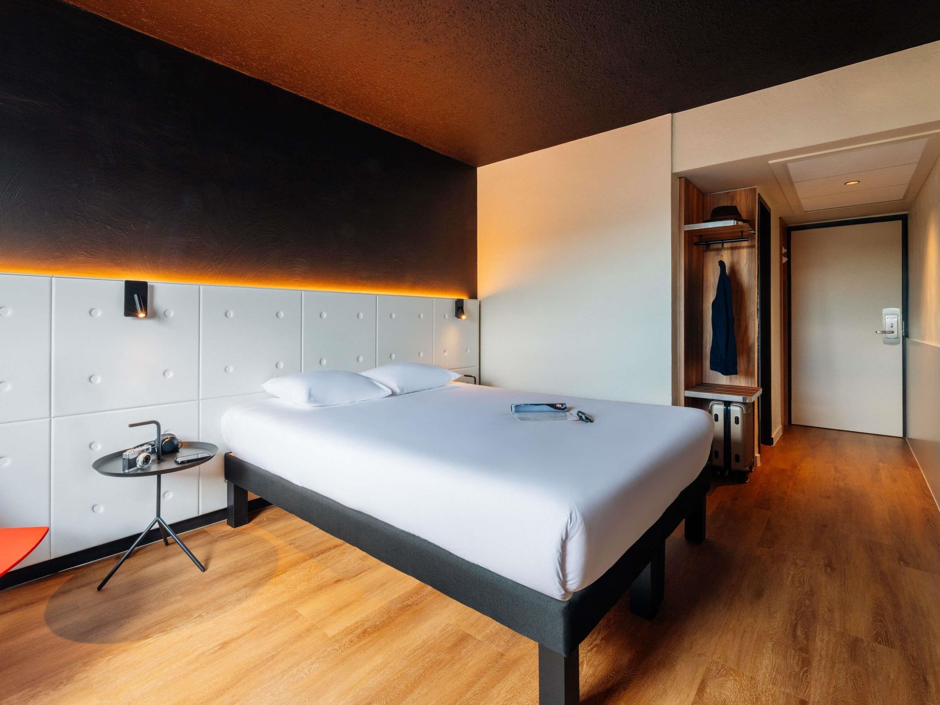 A modern hotel room with a large bed, a bedside table with a lamp, a wardrobe, and a shower area.