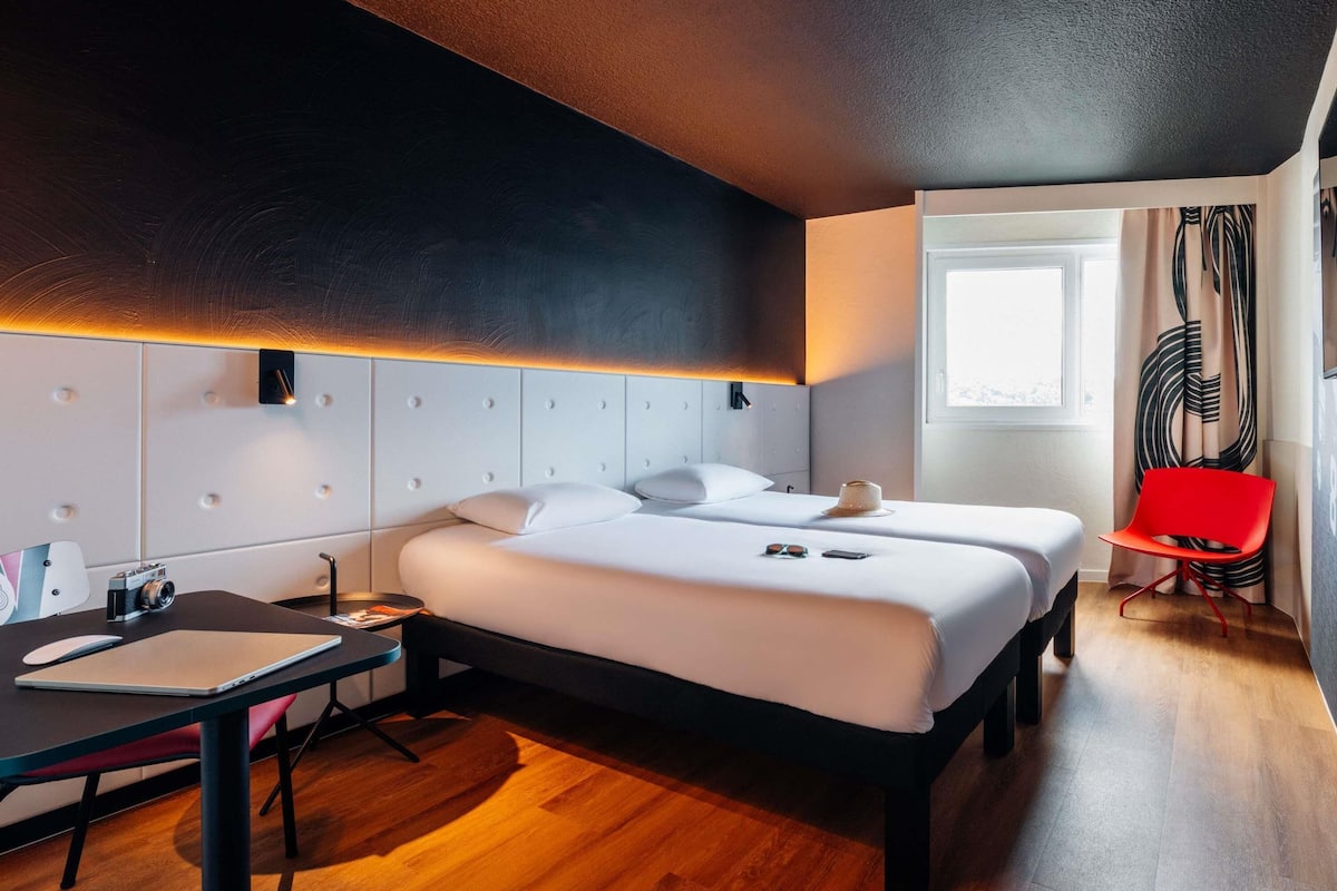 A modern hotel room with a large bed, a desk, a chair, and a window with curtains.