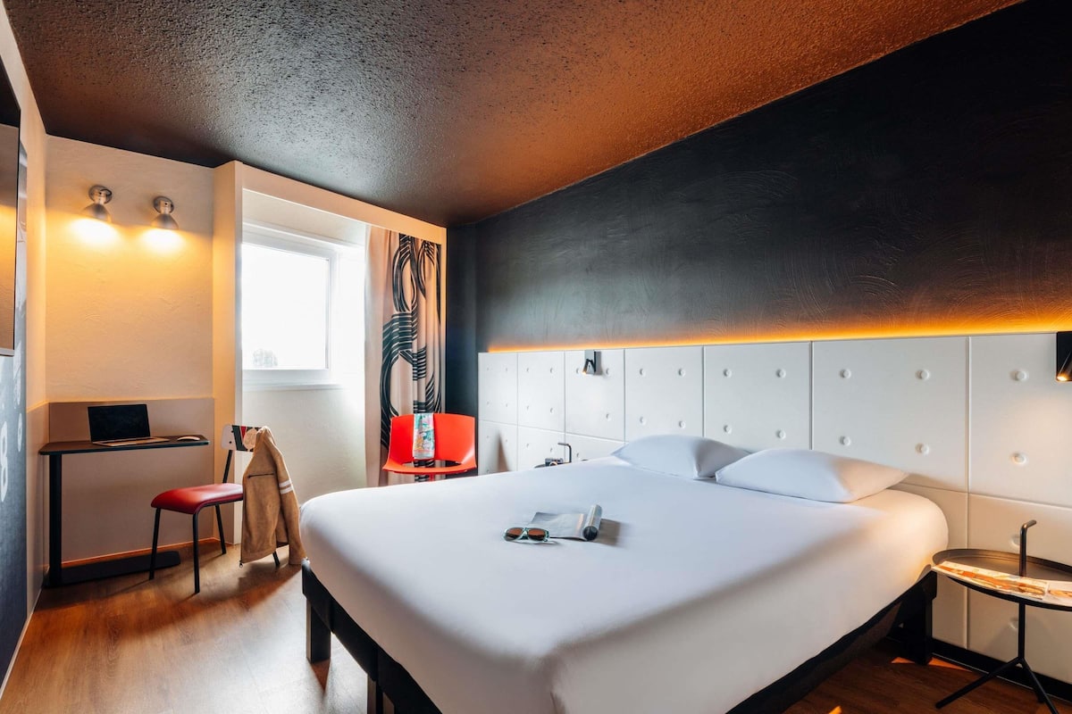 A modern hotel room with a large bed, a desk with a laptop, a chair, and a small table with a tray.