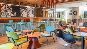 Uma área de bar moderna com teto de madeira, um aviso de 'Silêncio, por favor' e diversas opções de assento, incluindo mesas redondas e cadeiras coloridas.