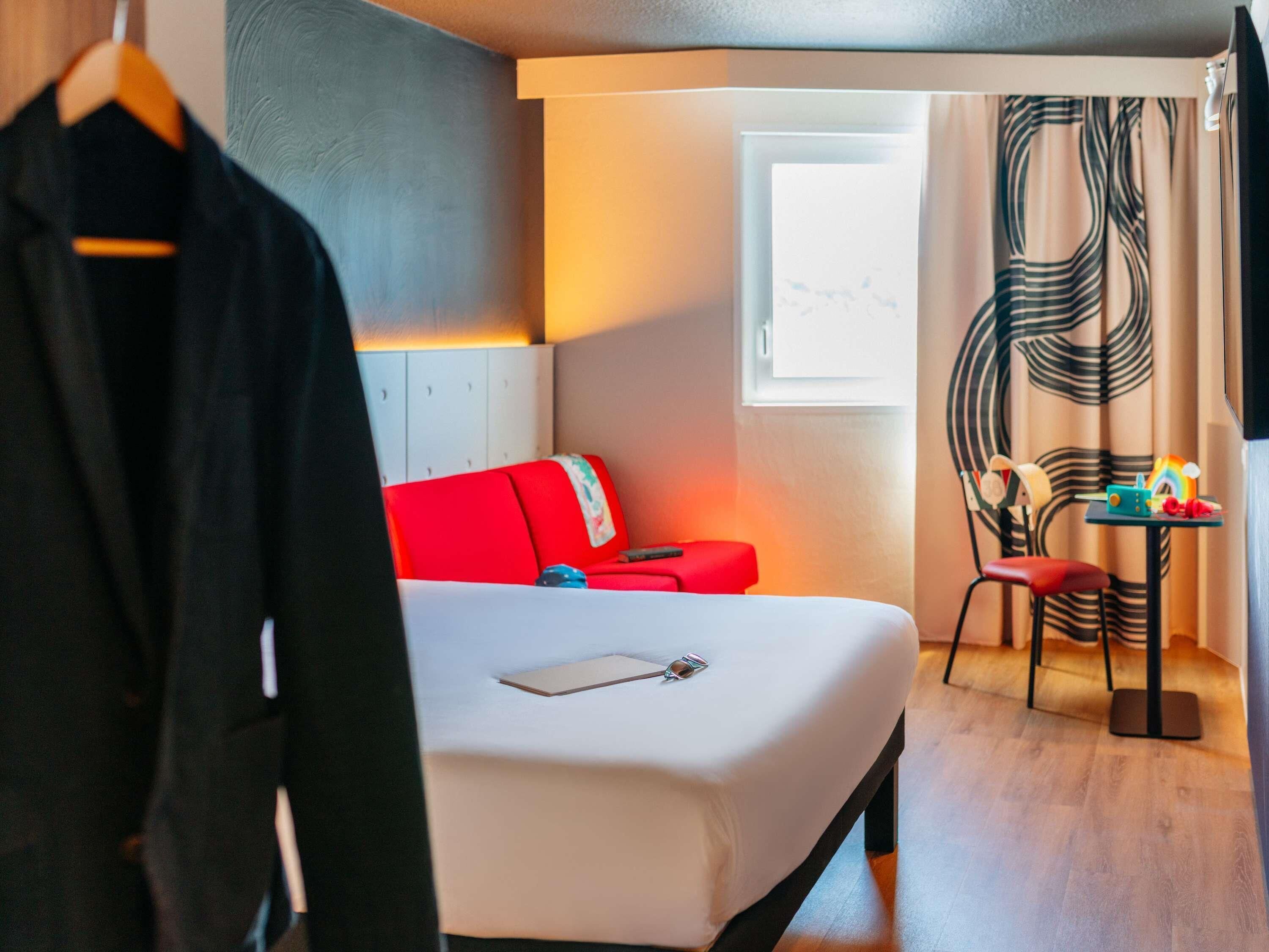 A hotel room with a bed, a red sofa, a small table with toys, and a window with curtains.