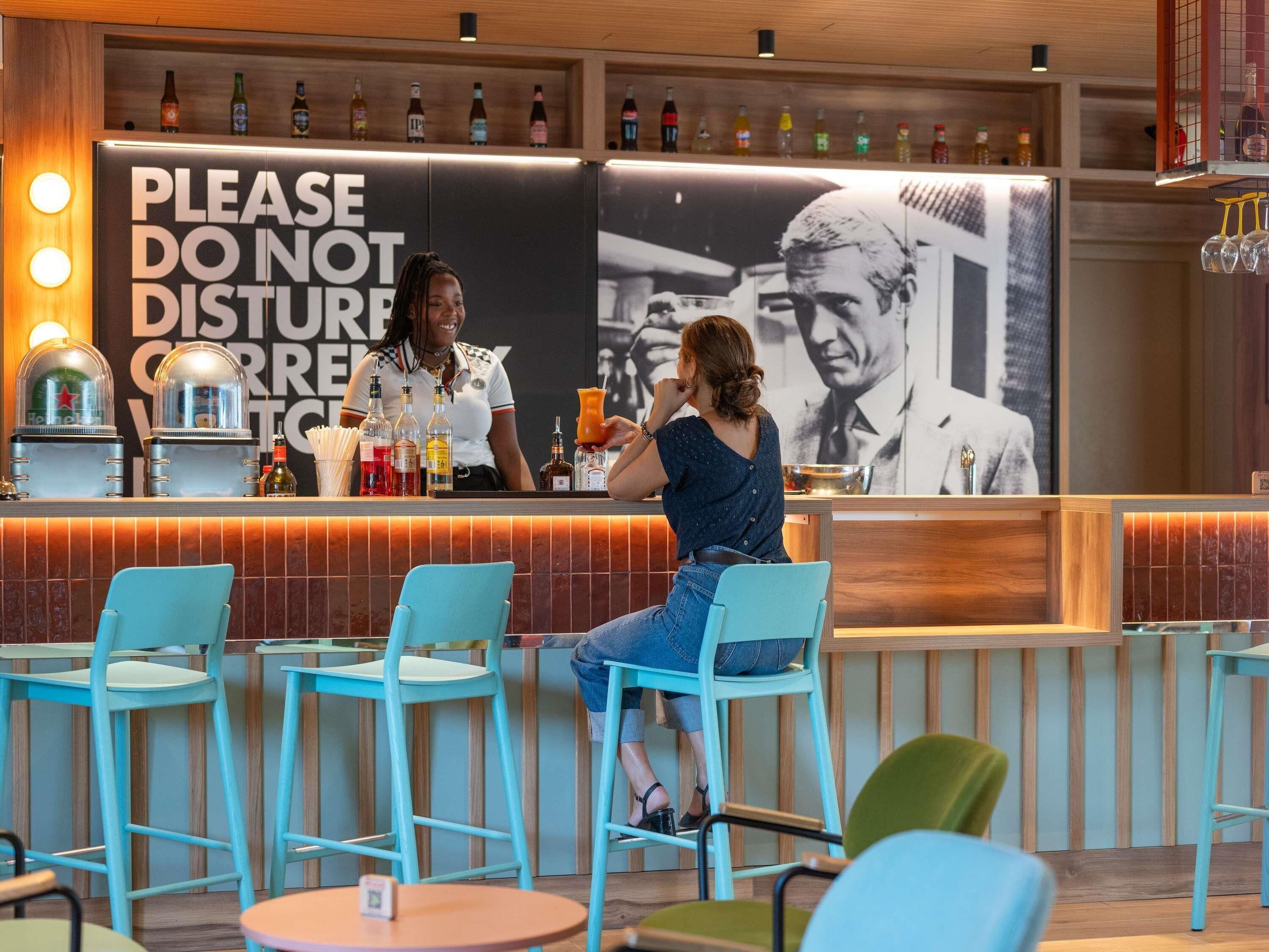 A bar area with a sign that reads 'PLEASE DO NOT DISTURB.' There are multiple bottles and glasses on the counter, and a person is seated at a table.