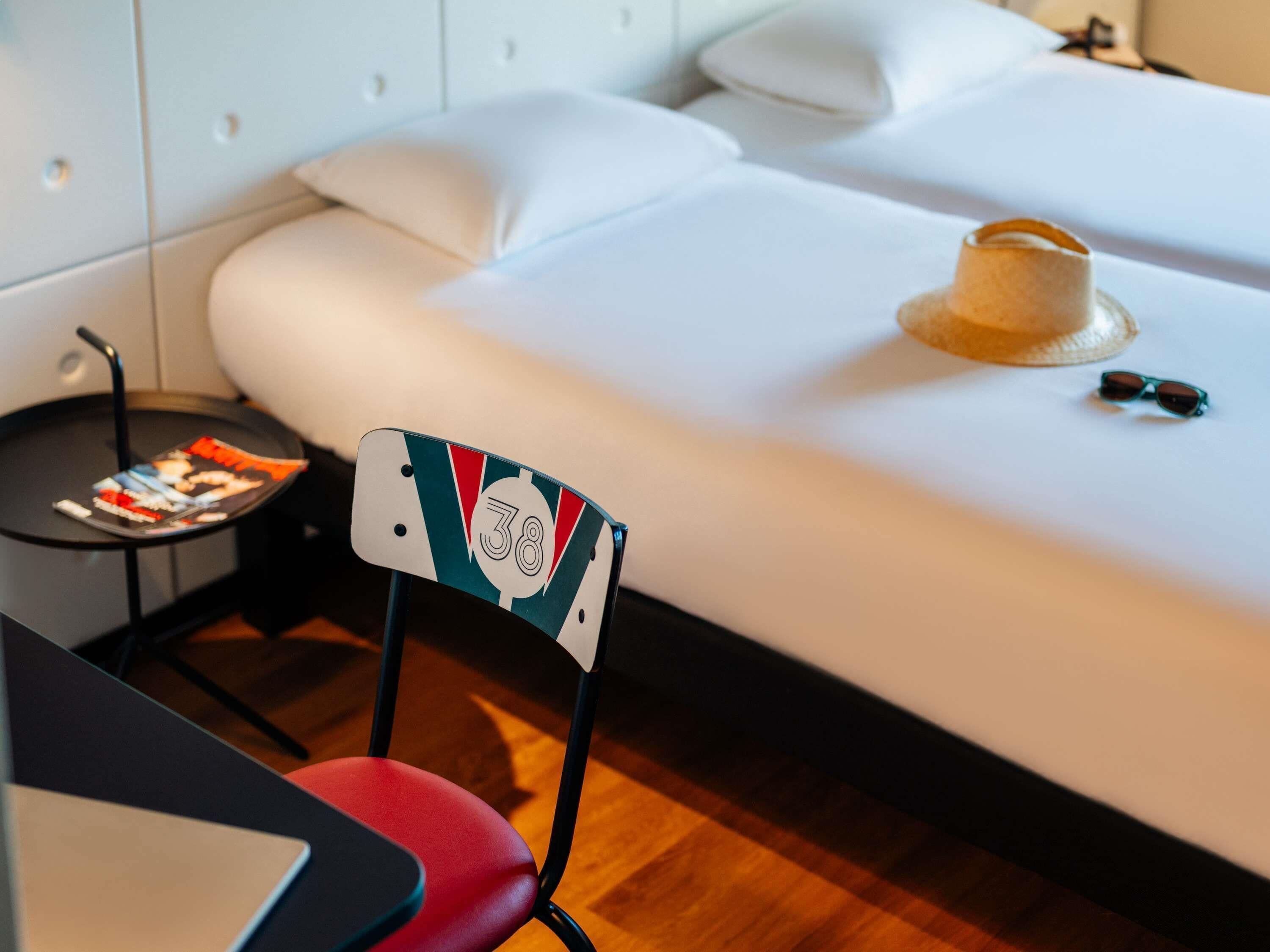 Una camera d'albergo con un letto, un tavolino con una rivista, una sedia con il simbolo del numero e un cappello sul letto.