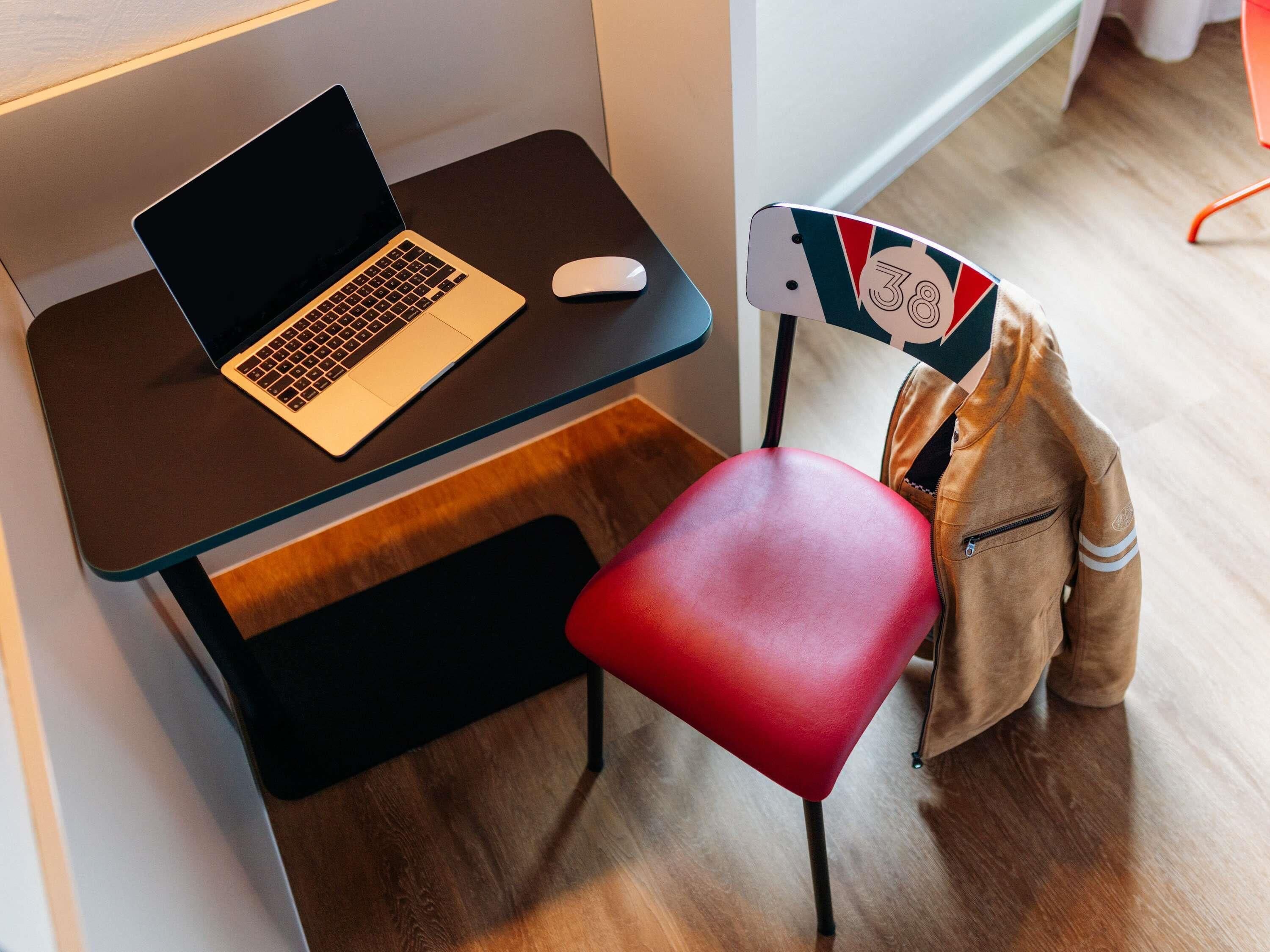 A laptop on a desk, a red chair, and a jacket hanging on a chair.