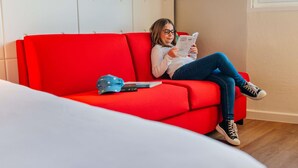 A person sitting on a red sofa in a room with a white headboard and a window. - ibis Styles Lyon Sud Vienne (Chasse-sur-Rhone)