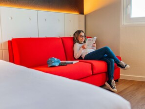 A person sitting on a red sofa in a room with a white headboard and a window. - ibis Styles Lyon Sud Vienne (Chasse-sur-Rhone)