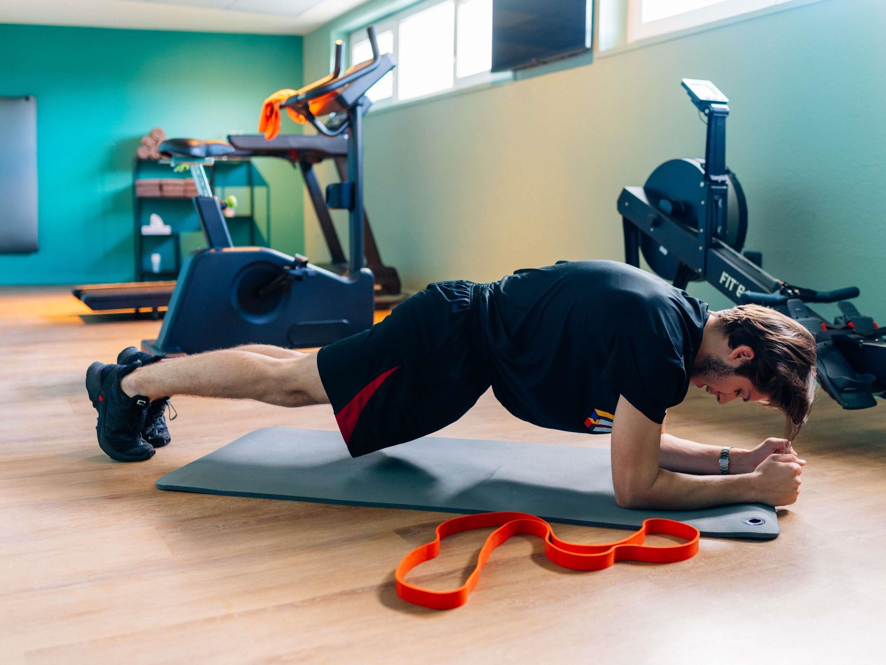 Une personne effectuant un exercice de planche sur un tapis dans une salle de sport, avec du matériel d'exercice en arrière-plan.