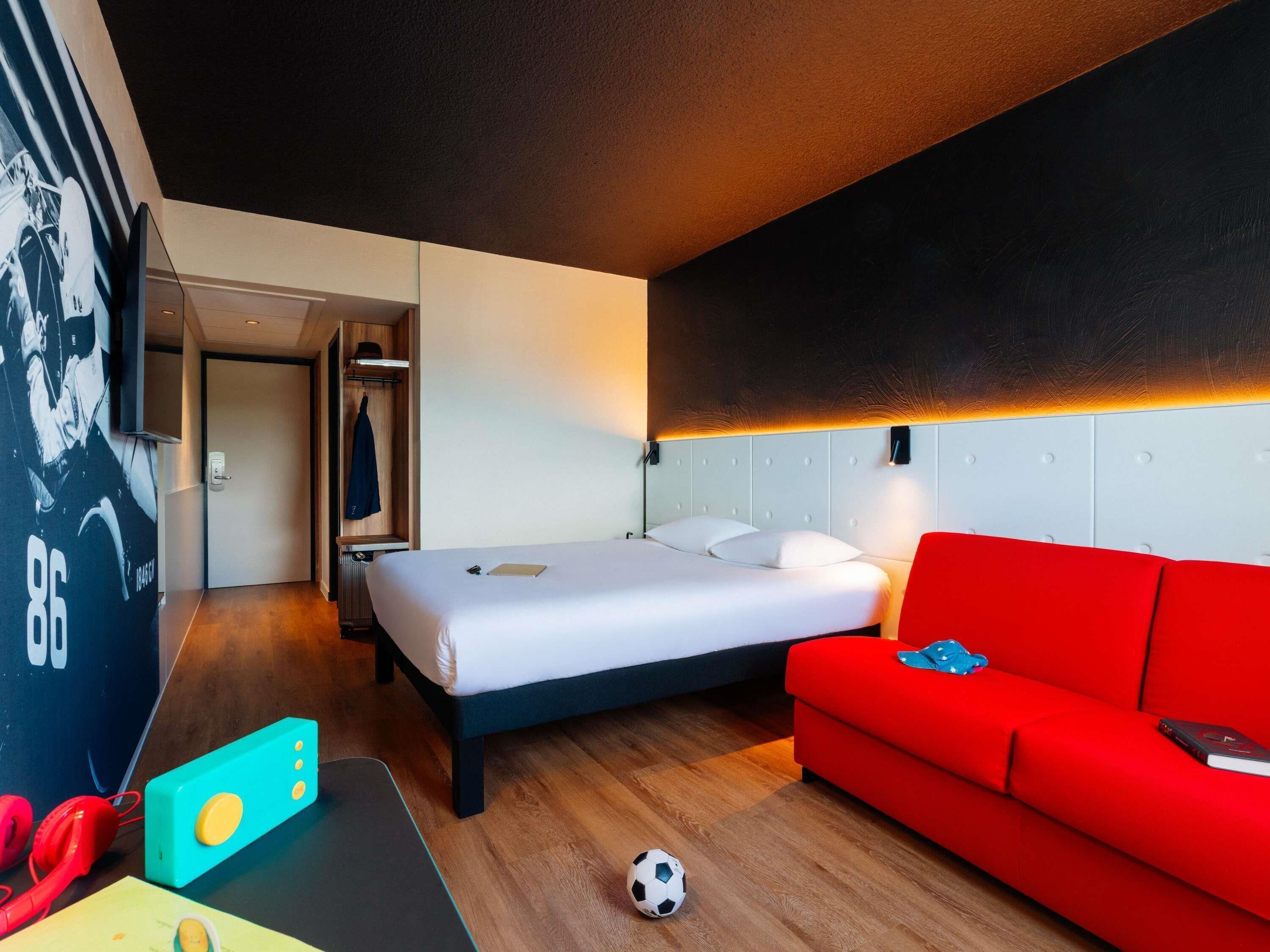 Une chambre d’hôtel avec un lit, un canapé rouge, un téléviseur fixé au mur et une porte donnant sur un couloir.
