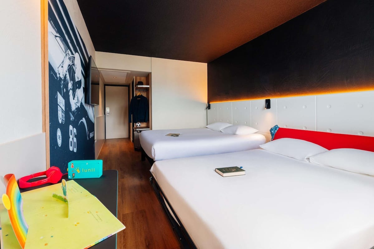 A hotel room with two beds, a desk with a laptop, a red and yellow toy, and a wall with a black and white photo.