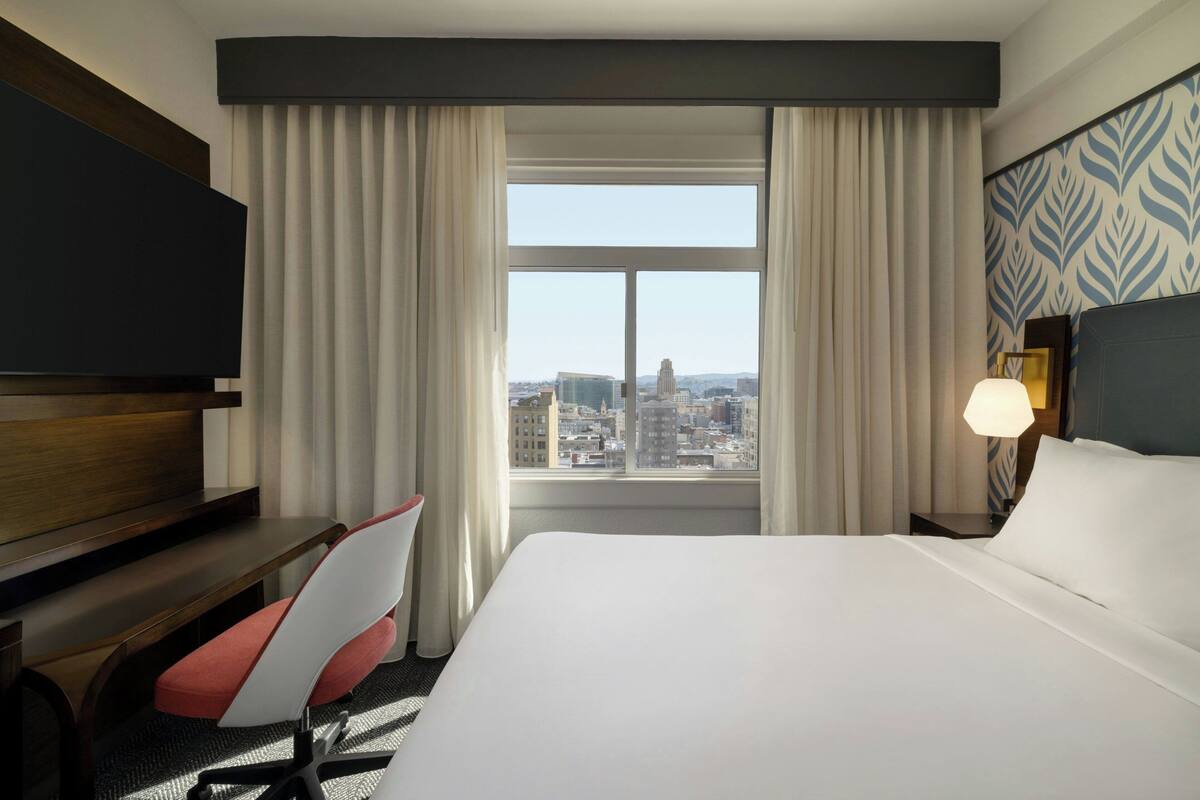 Habitación de hotel con una cama grande, un escritorio, una silla, un televisor y vistas a la ciudad desde la ventana.