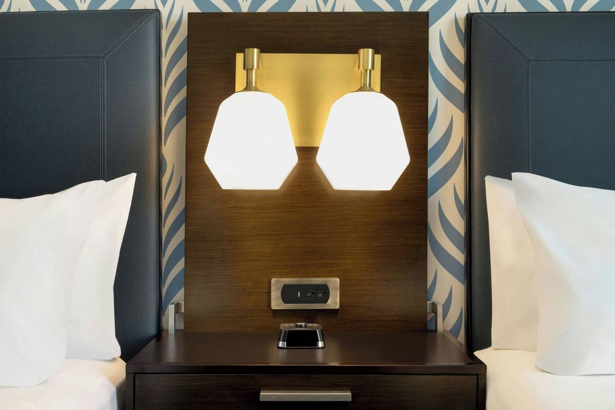 Una habitación de hotel con cabecera de madera, dos lámparas de noche, un reloj digital y una mesita de noche con cajón.