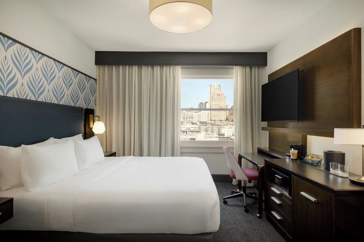 Habitación de hotel con una cama grande, un escritorio, una silla, un televisor y vistas de la ciudad.