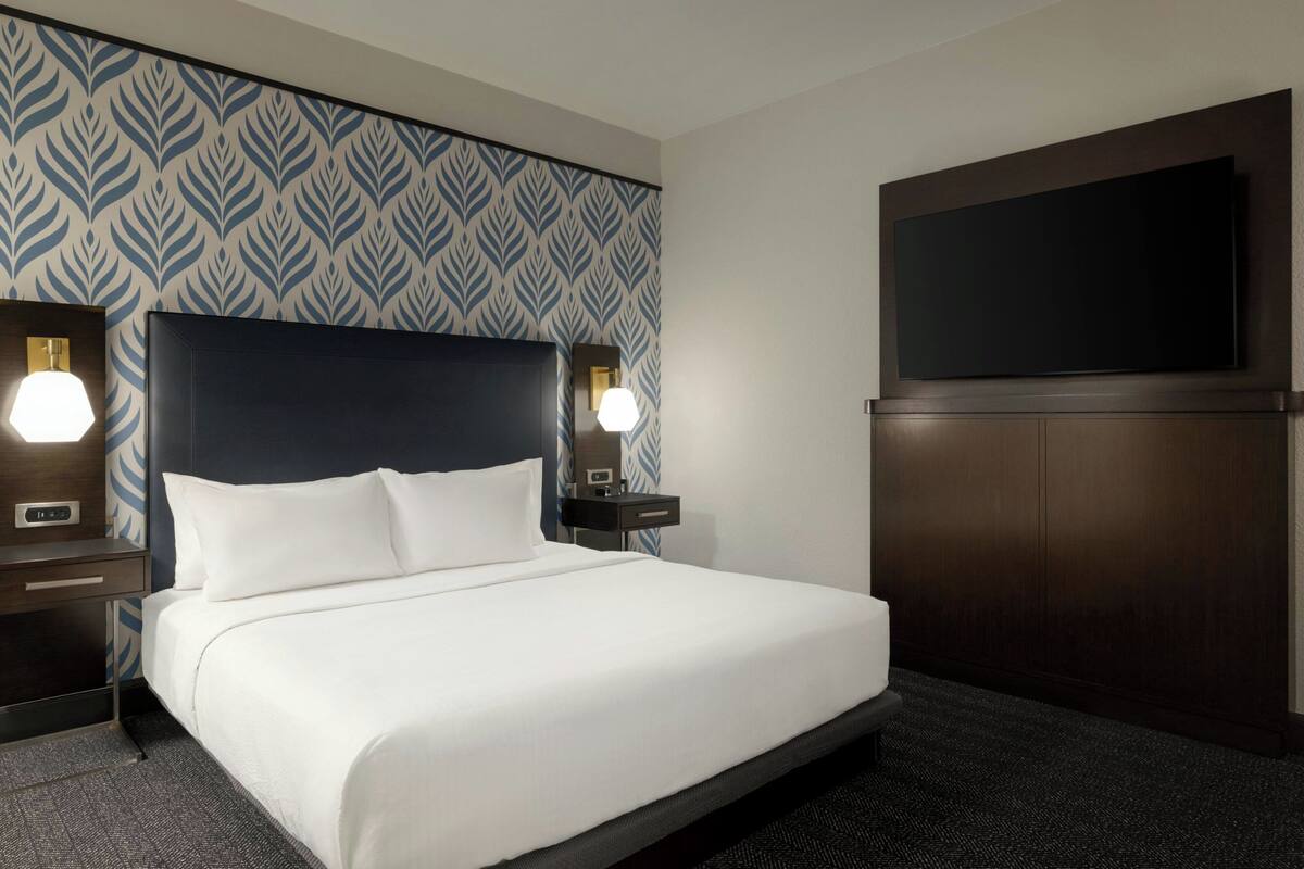 Habitación de hotel con una cama grande, dos mesitas de noche con lámparas, un televisor de pantalla plana montado en la pared y un papel tapiz estampado.