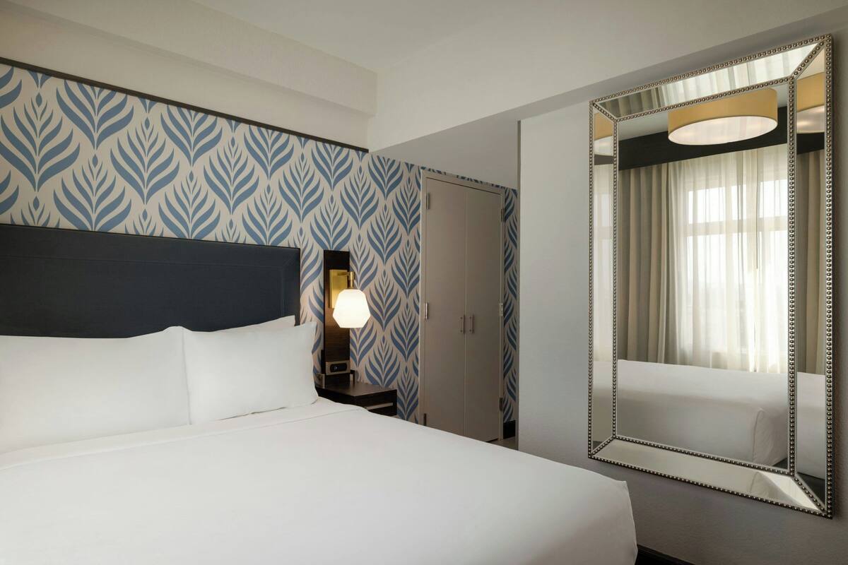 Una habitación de hotel con una cama grande, una mesita de noche con lámpara, un armario y un espejo con marco decorativo.